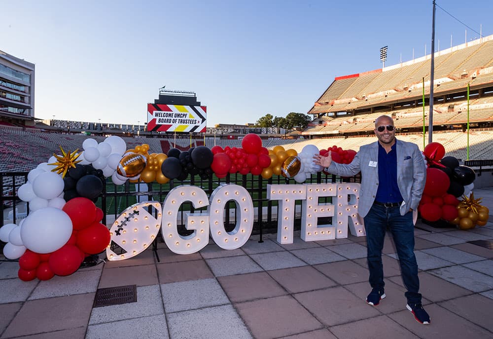 Trustee Mike Luzio posing in front of a light up Go Terps sign and balloons.