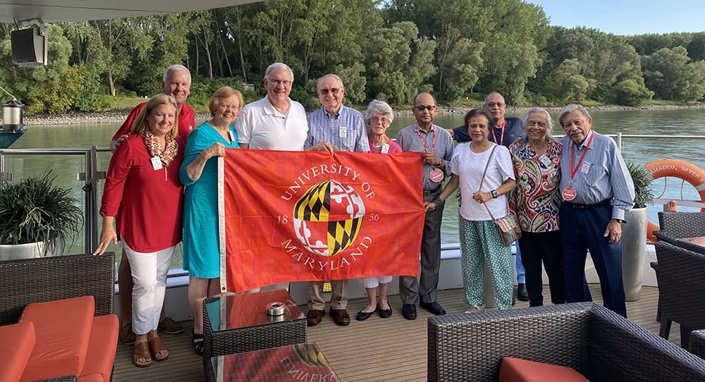 A group of Terp alumni posing together on a river cruise boat while holding a University of Maryland flag