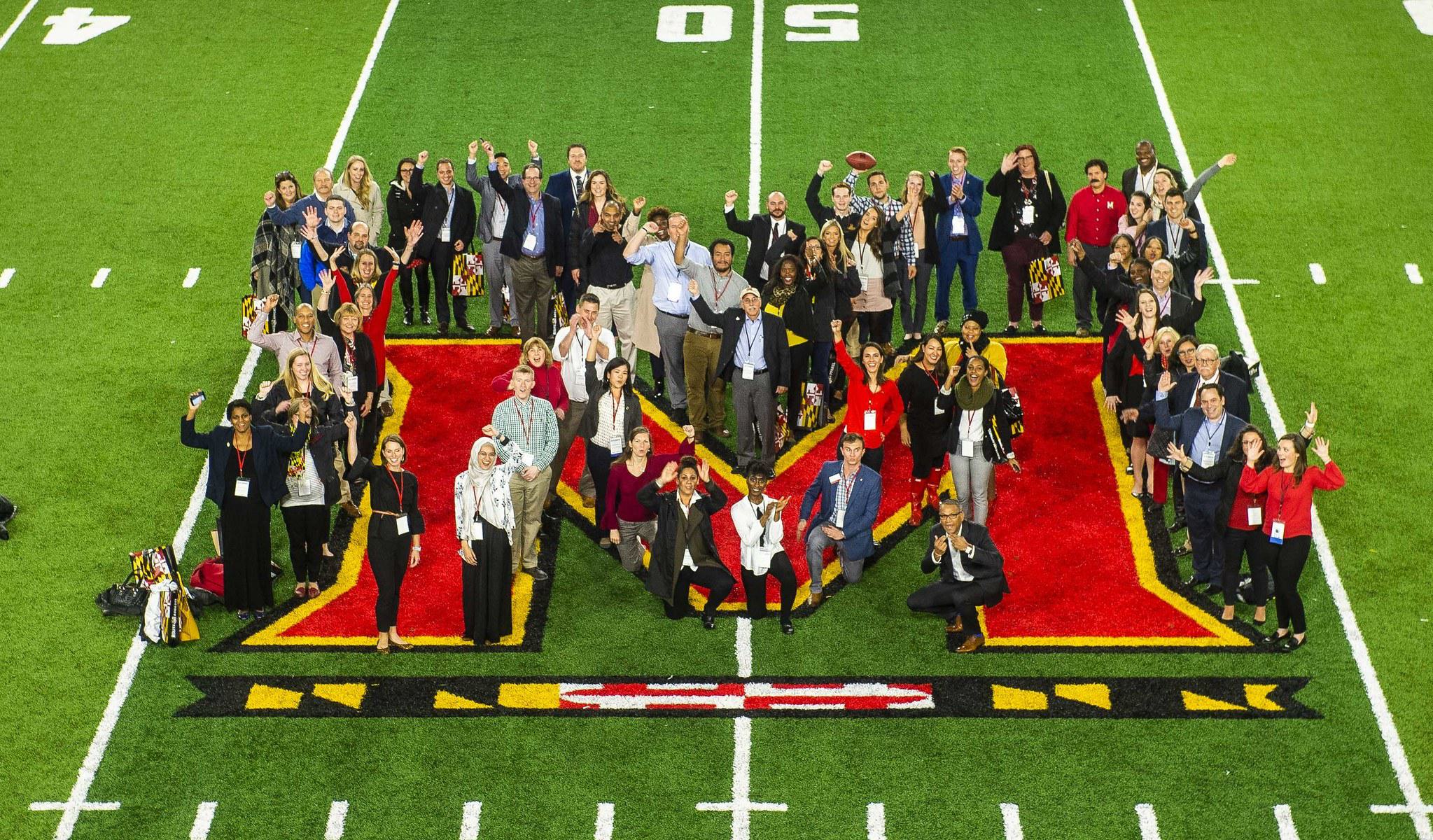 A group Alumni honorees pose outlining the Maryland M at the center-line of Capital One field. The camera is positioned high in the stadium bleachers to get a wide view of everyone