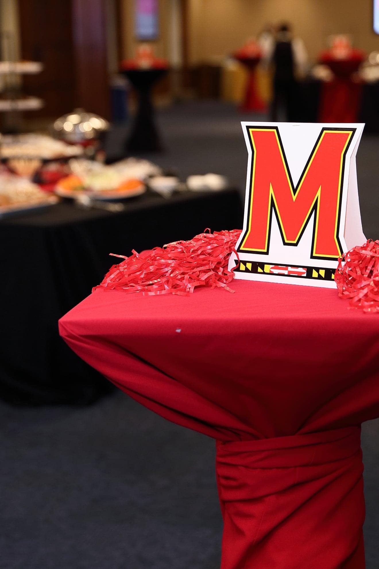 The Maryland M center piece on a high-top table draped in a Maryland Red cloth. out of focus in the background a table of refreshments for an event