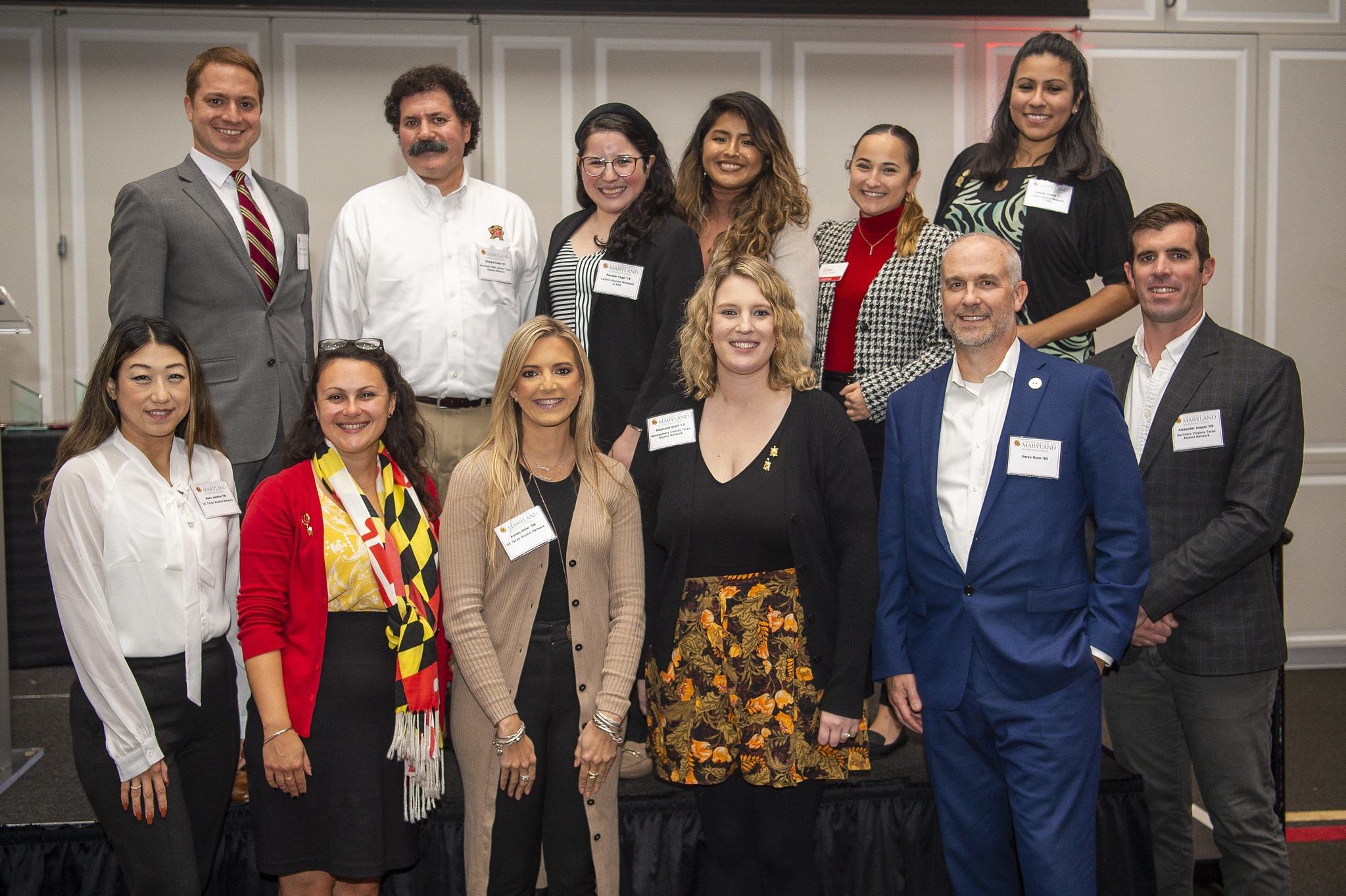 Alumni Honorees pose in two rows, one row on the event stage, the second in front of it. everyone is smiling for the camera