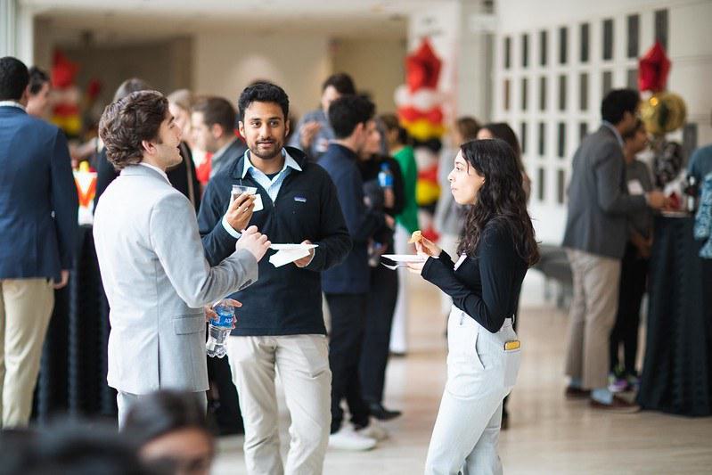 Several attendees of a University event stand facing each-other, plates and water-bottles in hand, discussing relevant topics
