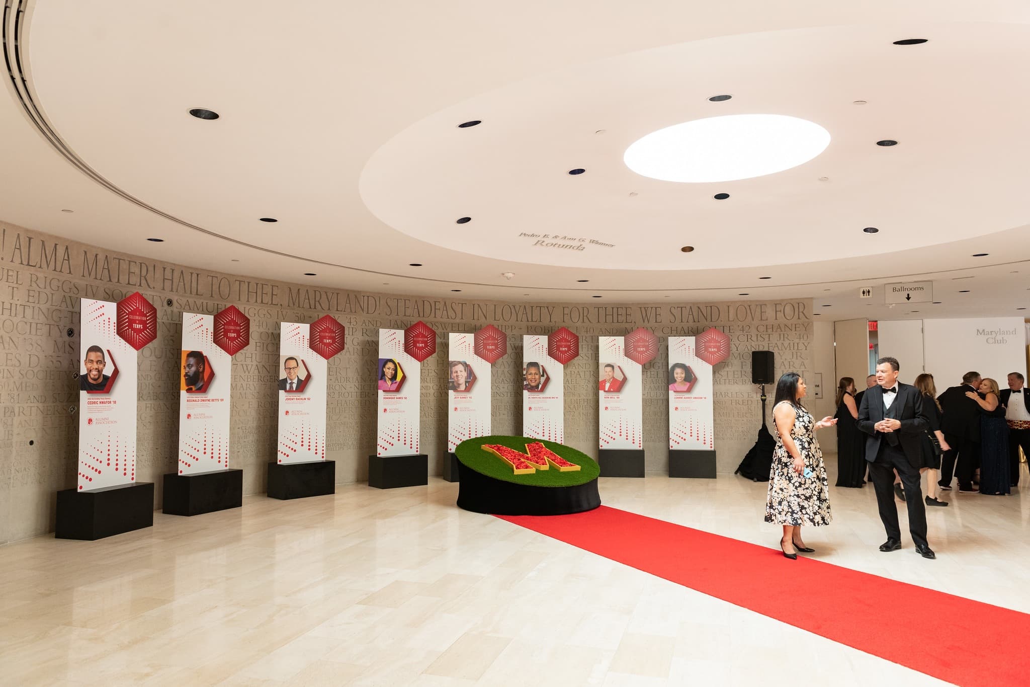 The Samuel Riggs IV Alumni Center rounded lobby, decorated with a red carpet over a clean white marble floor. Tall banners each featuring an award winner of the festivities lines the wall.