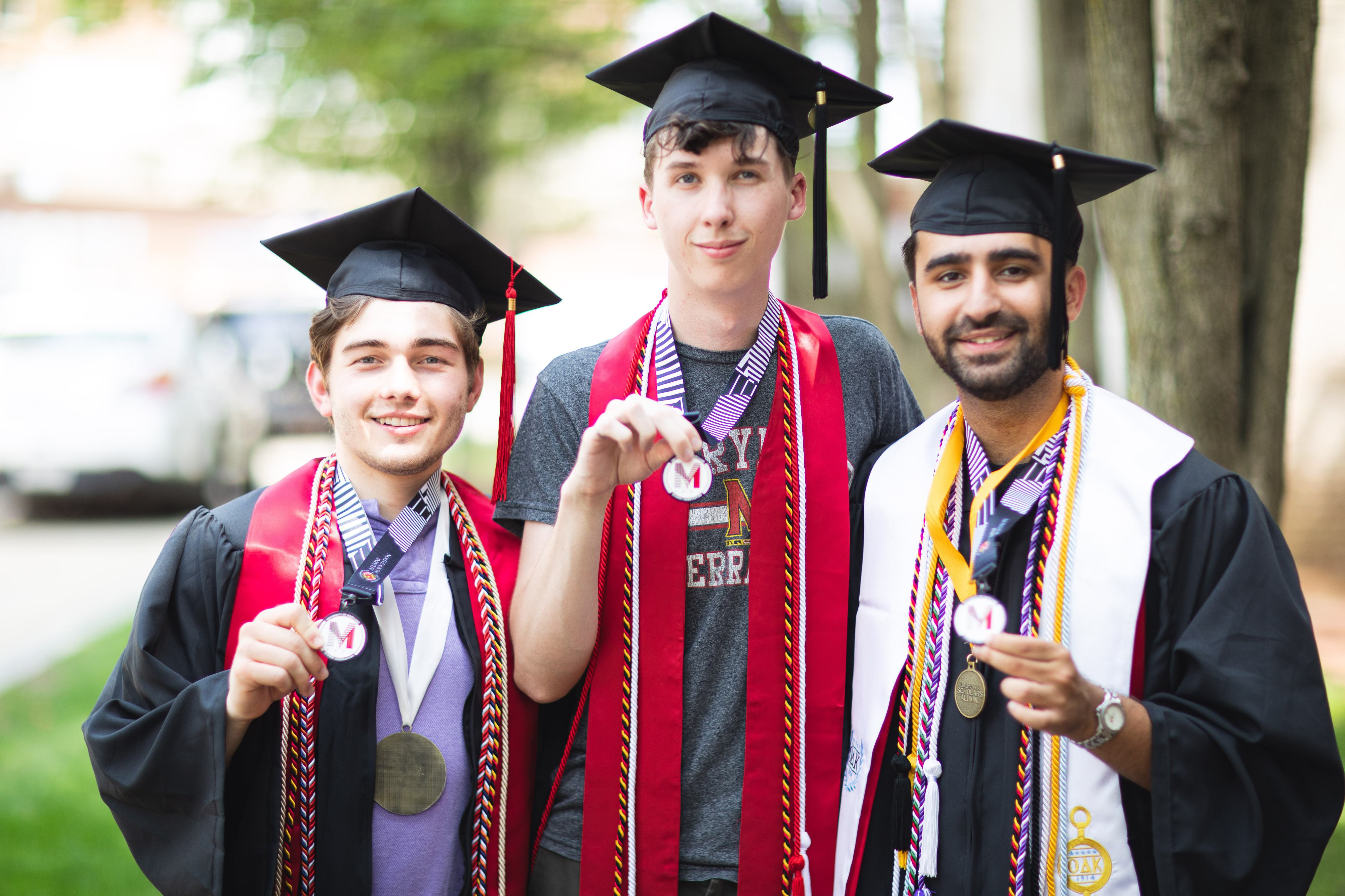 Three Recent Graduates in full graduation regalia proudly present their M Book Lanyards for completing the UMD Bucket List
