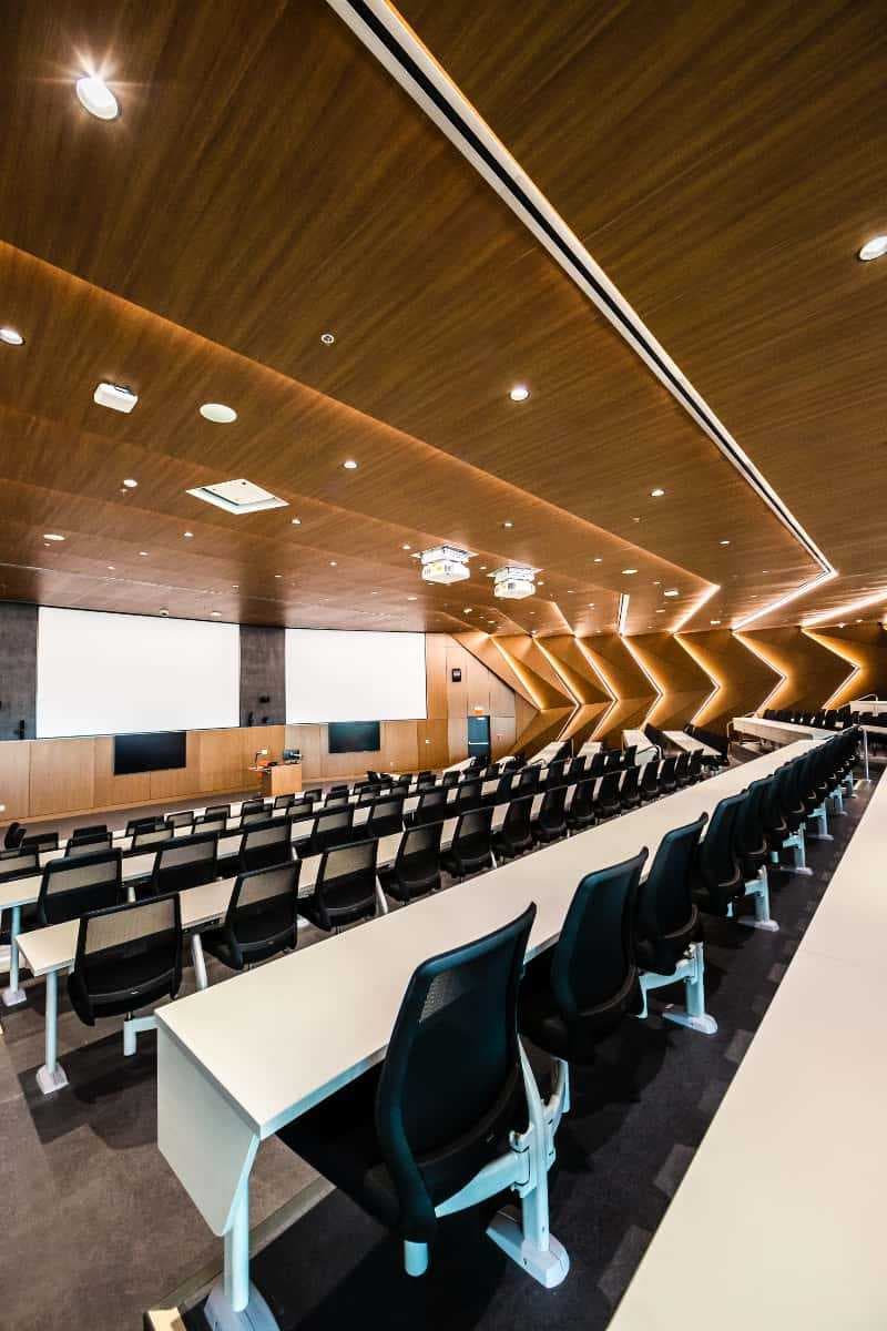 Modern Auditorium with in-set lighting along bright wood-panel walls. Comfortable seats behind long bench tables rack down towards the front of the room.