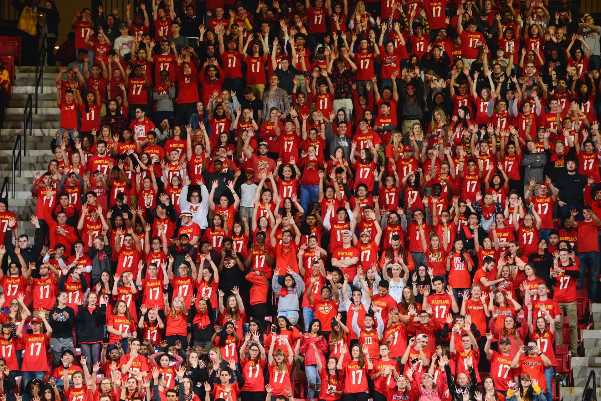 Wide shot of fans at a University of Maryland Basketball game standing and cheering, everyone is wearing some for of Maryland Red attire