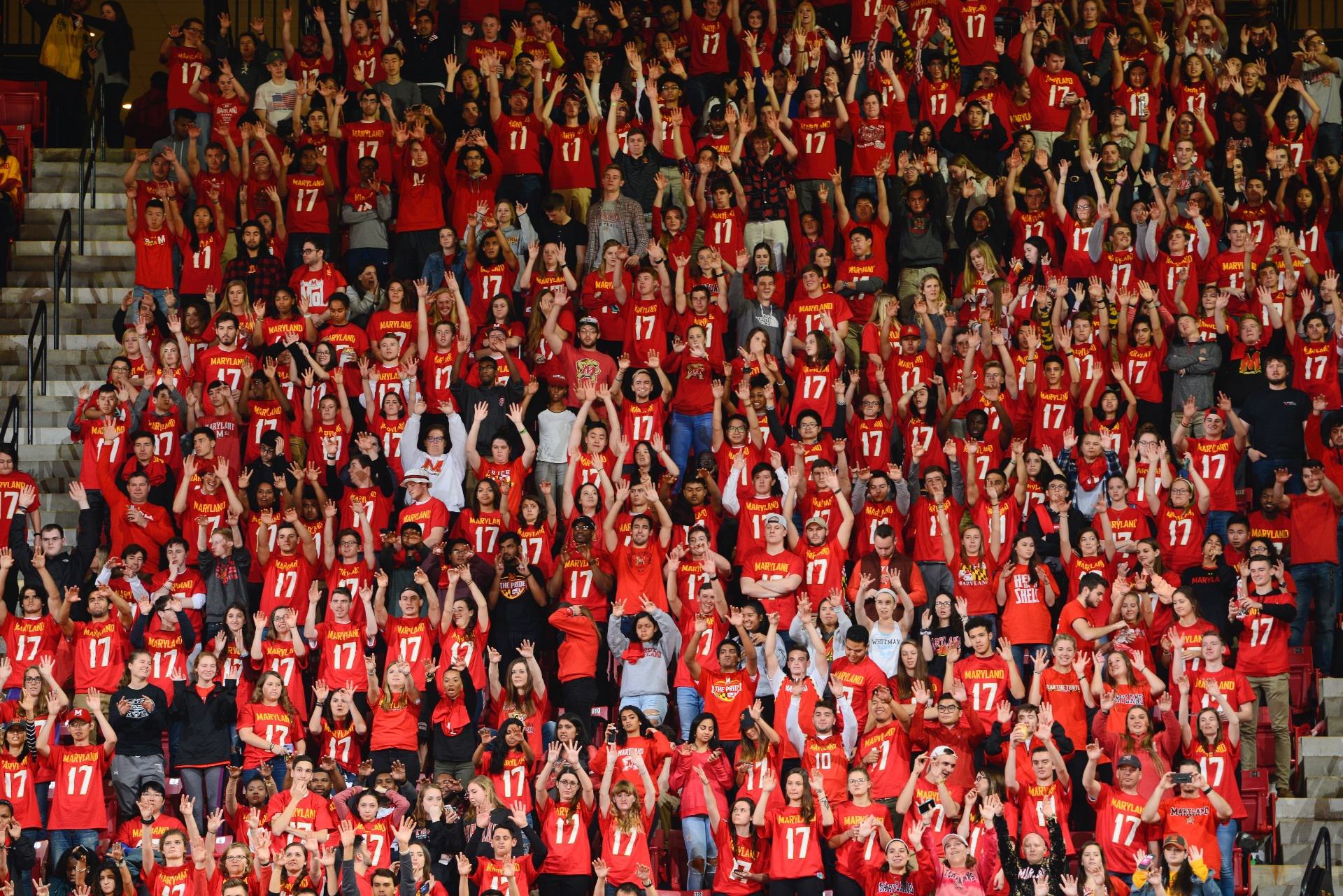 Wide shot of fans at a University of Maryland Basketball game standing and cheering, everyone is wearing some for of Maryland Red attire